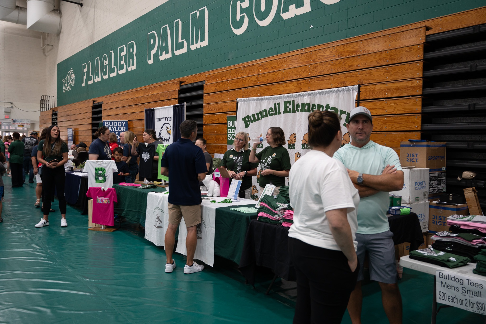 People are gathered at tables inside a gymnasium for a school event Signs for "Buddy Taylor Middle School" and "Bunnell Elementary" are visible Some stand while others browse or converse The gym walls are green with "FLAGLER PALM COAST" written prominently