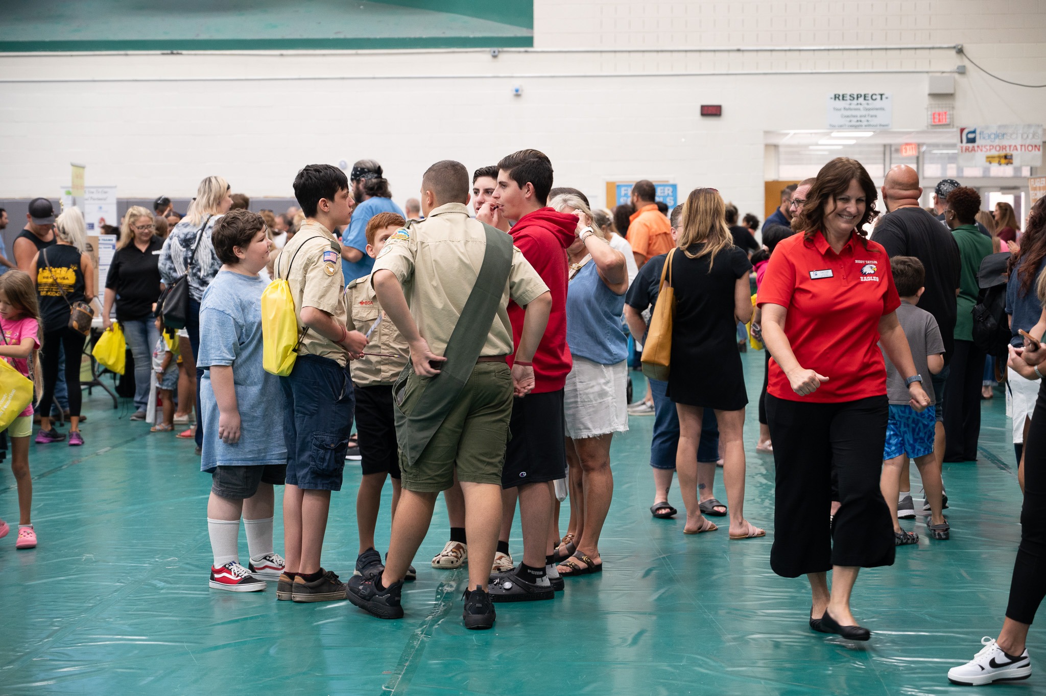 People are gathered in a large indoor space A group of youths in scouting uniforms converse in the center A woman in a red shirt interacts with participants to the right Attendees are engaged in various activities, and several informational booths are visible in the background