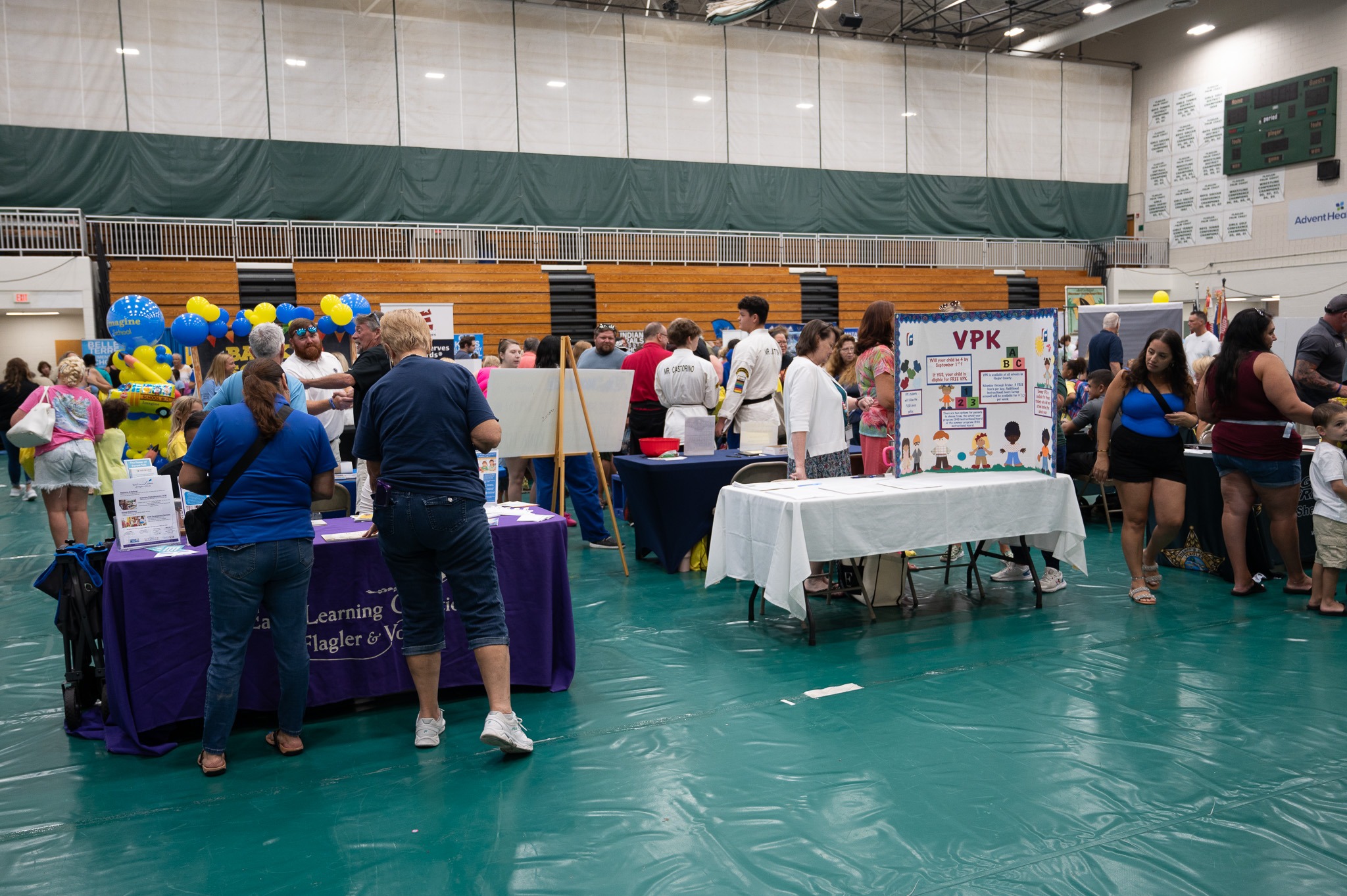 A busy indoor event with numerous people interacting with various booths in a large gymnasium Tables are covered with informational materials and signs, including one for VPK (Voluntary Prekindergarten) Balloons and posters add to the colorful atmosphere