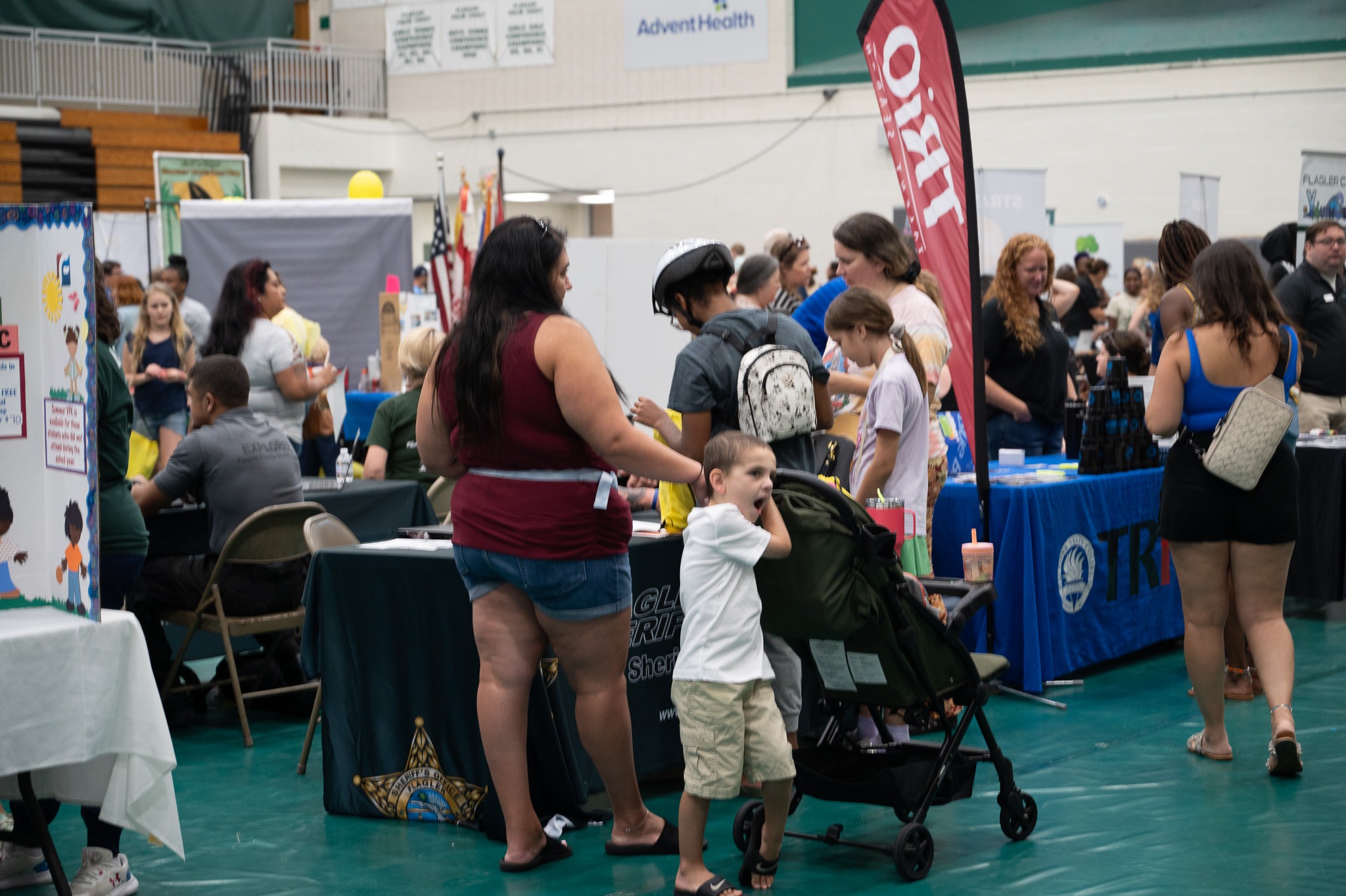 A busy indoor fair with people visiting different booths A woman with long dark hair stands next to a man in a black shirt while a child in a white shirt pushes a green stroller Several banners, tables, and displays are visible in the background