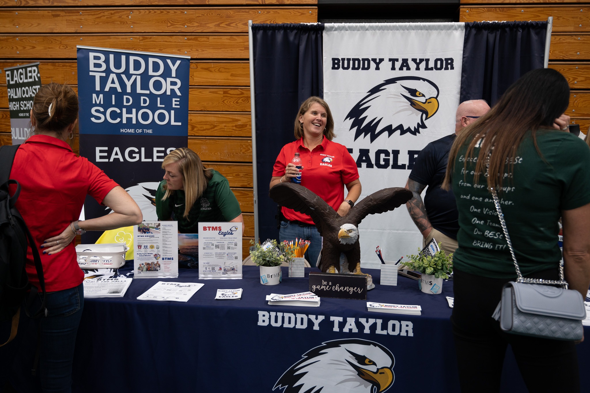 People at a booth for Buddy Taylor Middle School with a table covered in materials and decorations, including an eagle figurine A banner in the background features the school's logo, and a woman wearing a red shirt is smiling and engaging with attendees