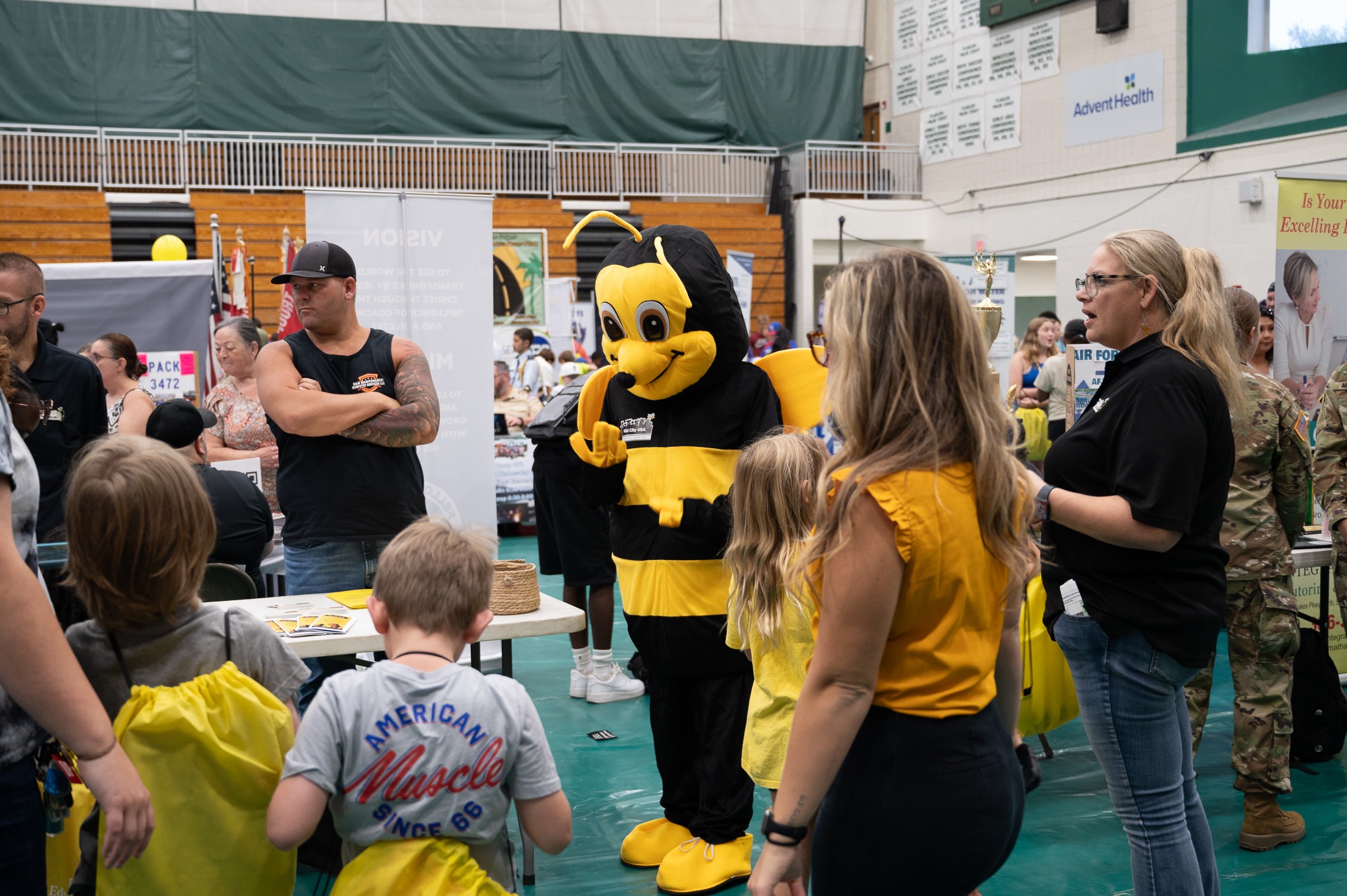 A brightly colored bee mascot interacts with children and adults at a crowded indoor event People stand around booths and tables decorated with various informational materials and giveaways The venue is a gymnasium with a green floor and white walls