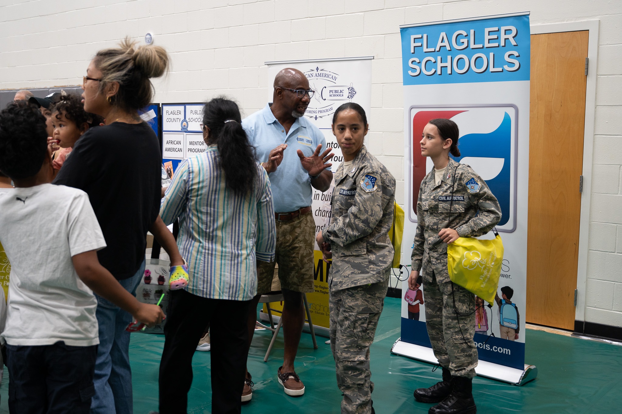 A group of people is gathered at an event in a gymnasium Two individuals in military uniforms stand near a "Flagler Schools" banner Others, including a man in a blue shirt and shorts, engage in conversation Children and adults are present, all interacting