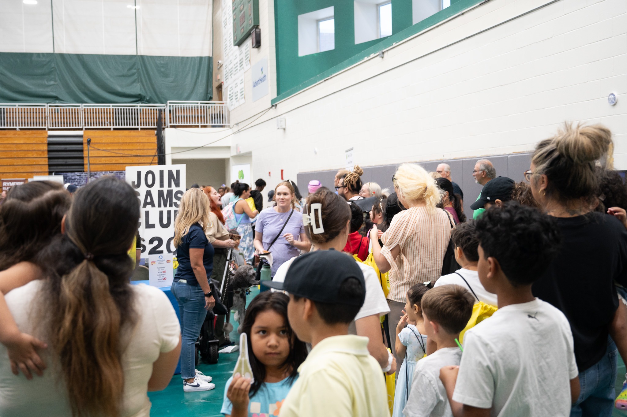 A gymnasium filled with a diverse crowd of adults and children A sign reading "Join AMAMS Club" and "$" is visible on the left Some people are standing in line, and others are milling around, engaging in various activities The atmosphere appears lively and bustling