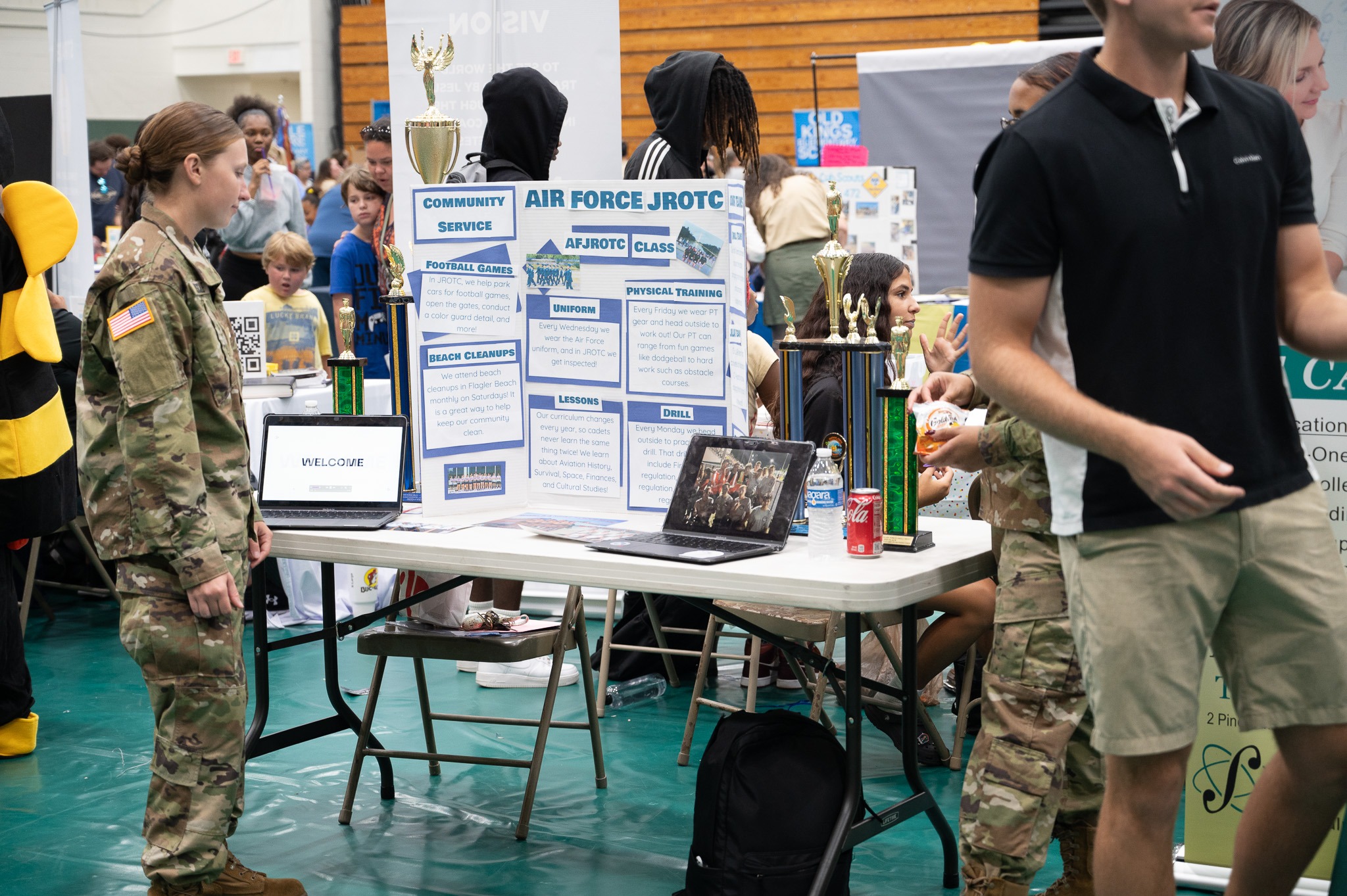 A busy indoor event displays an Air Force JROTC booth, featuring informational posters, laptops, and trophies Several individuals, including a uniformed service member, engage with the booth while others walk by in the background