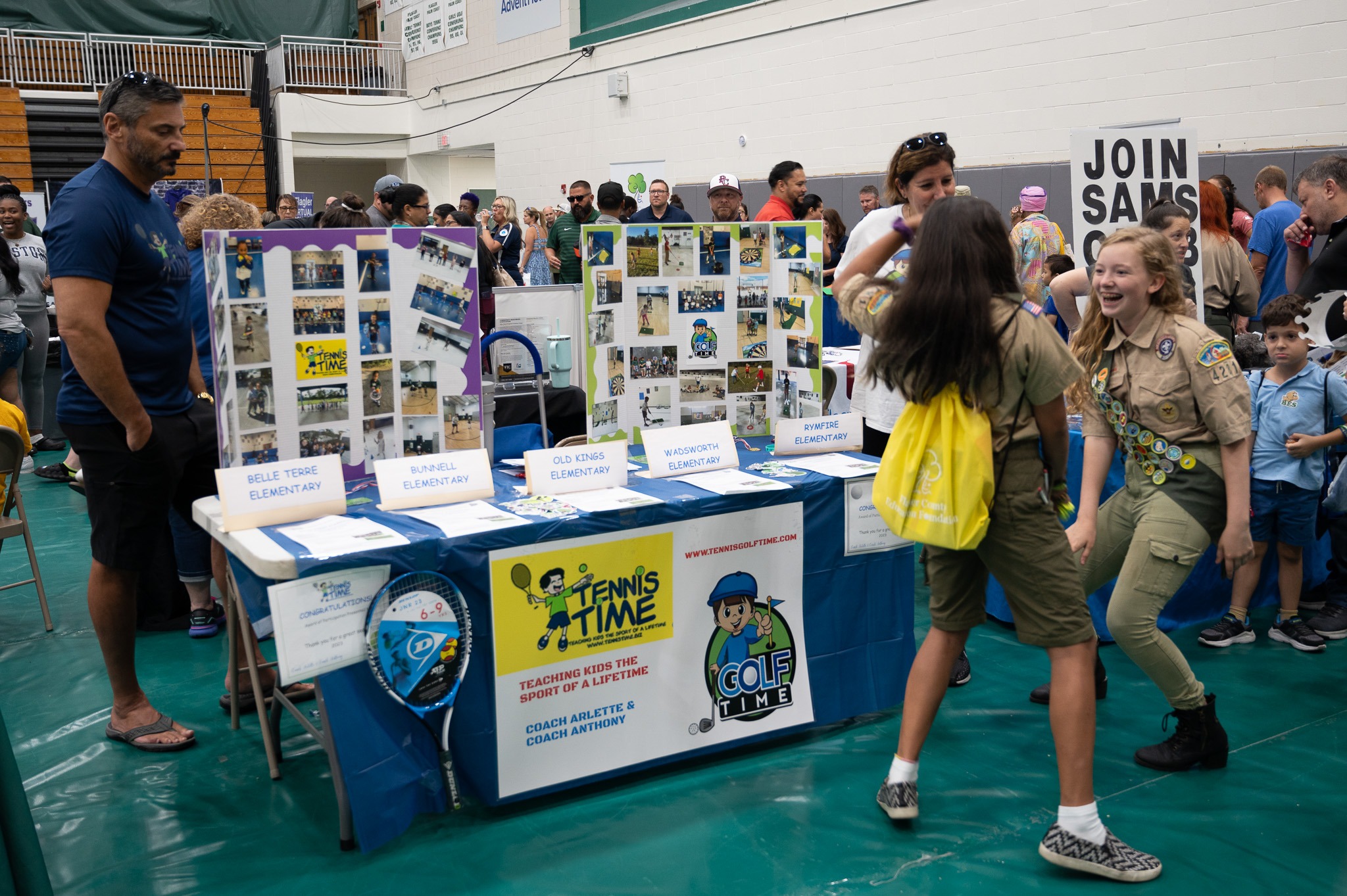 People visiting an exhibition booth in a gym The booth displays sports related information with posters for "Tennis Time" and "Golf Time" programs Two scouts are smiling and interacting with visitors Other people are looking around and conversing with one another