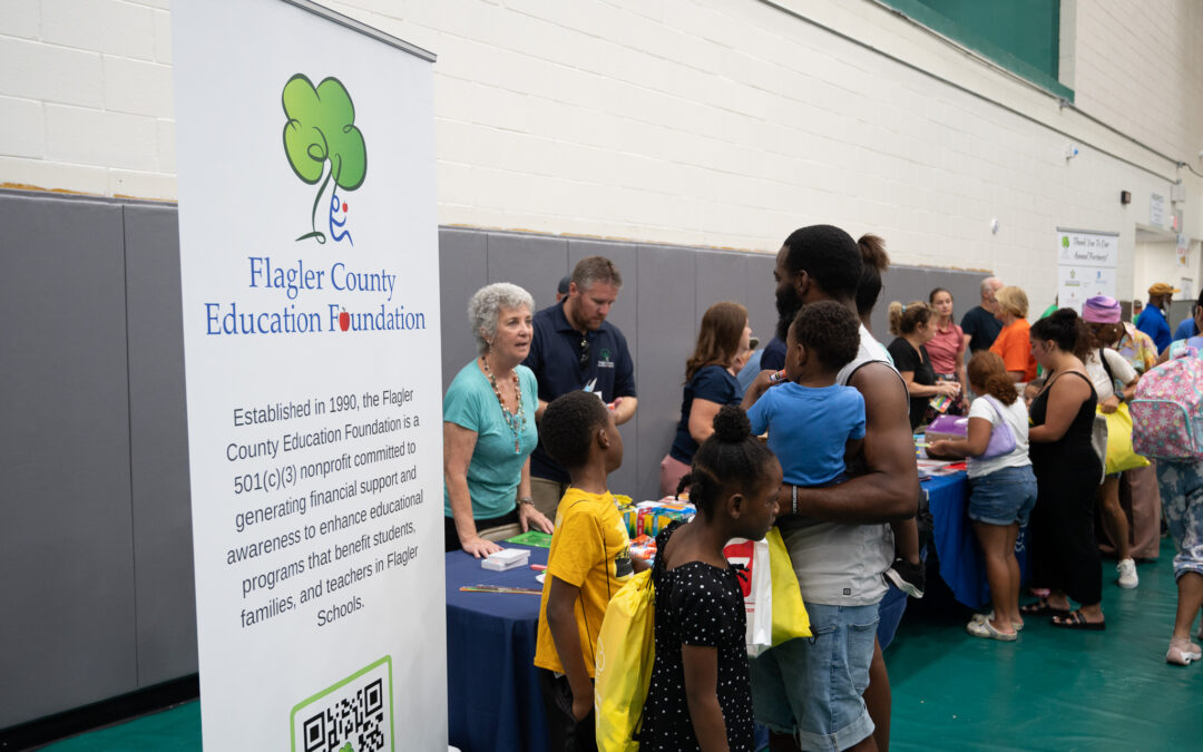 People gather at a community event in a gymnasium, interacting with representatives from various organizations A man and children stand at a booth for the Flagler County Education Foundation, where a banner provides information about the foundation’s mission