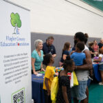 People gather at a community event in a gymnasium, interacting with representatives from various organizations A man and children stand at a booth for the Flagler County Education Foundation, where a banner provides information about the foundation’s mission