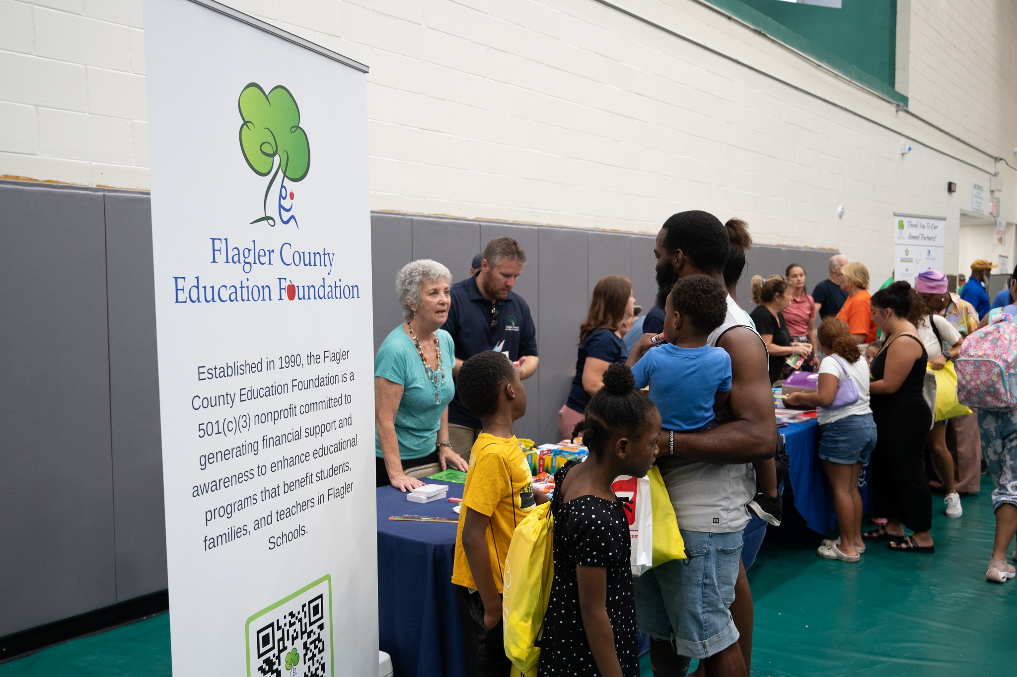 People gather at a community event in a gymnasium, interacting with representatives from various organizations A man and children stand at a booth for the Flagler County Education Foundation, where a banner provides information about the foundation’s mission