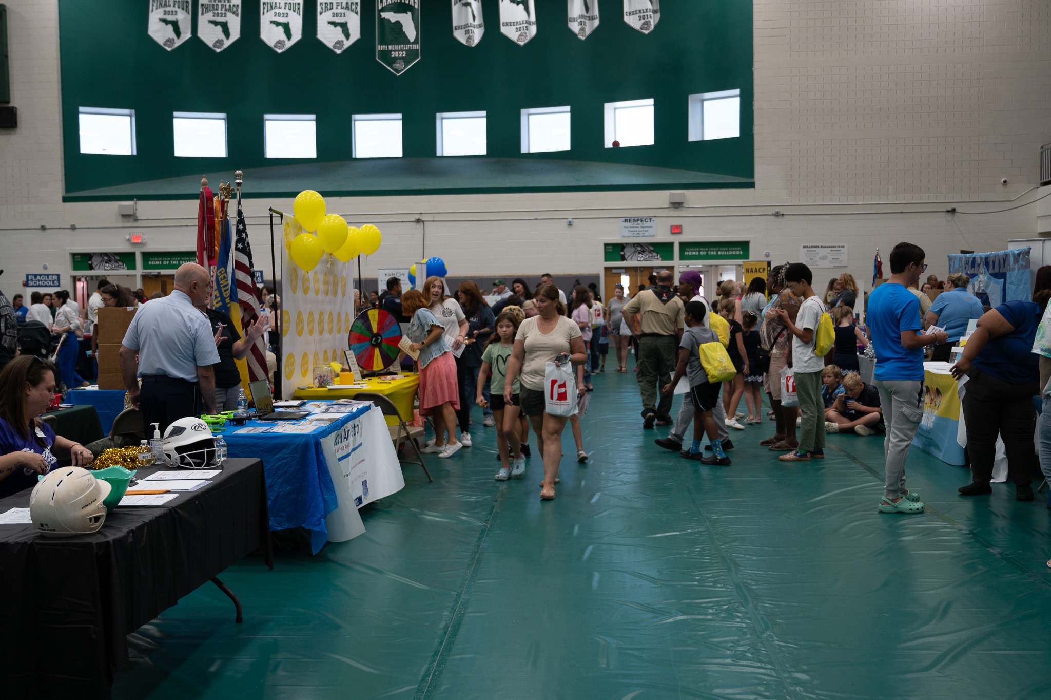 A bustling community fair takes place in a gymnasium with various tables and booths set up along the walls People of all ages, including children, are exploring the booths, which feature items like balloons and informational displays under green and white banners