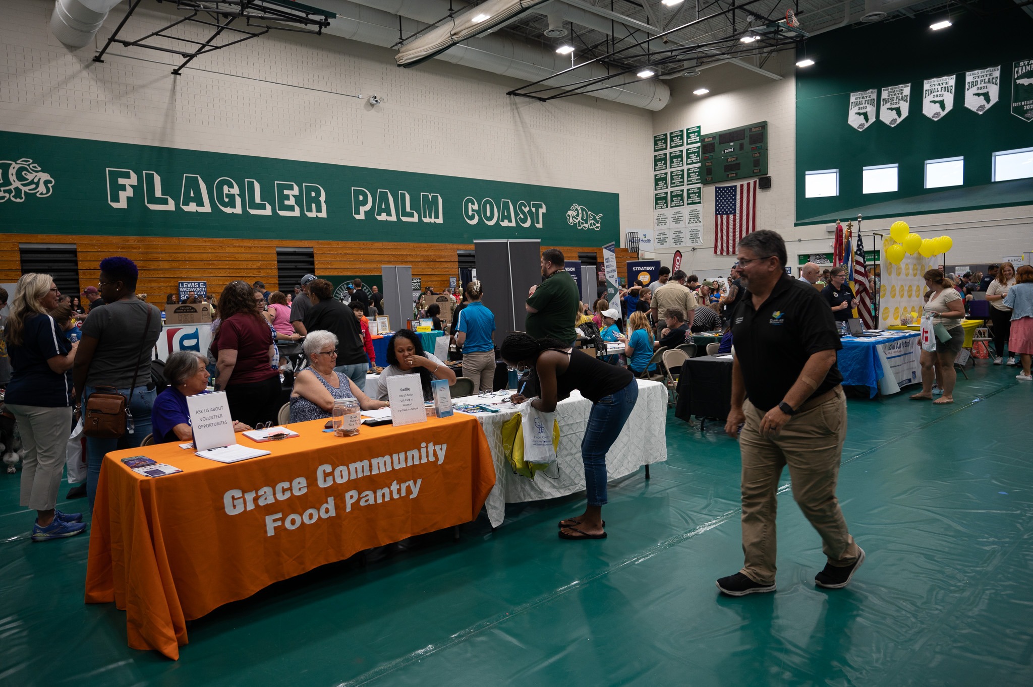 Community members engage with various booths at a local fair held in a gymnasium One prominent booth is for Grace Community Food Pantry with an orange tablecloth The background shows banners and the words "Flagler Palm Coast" on the wall