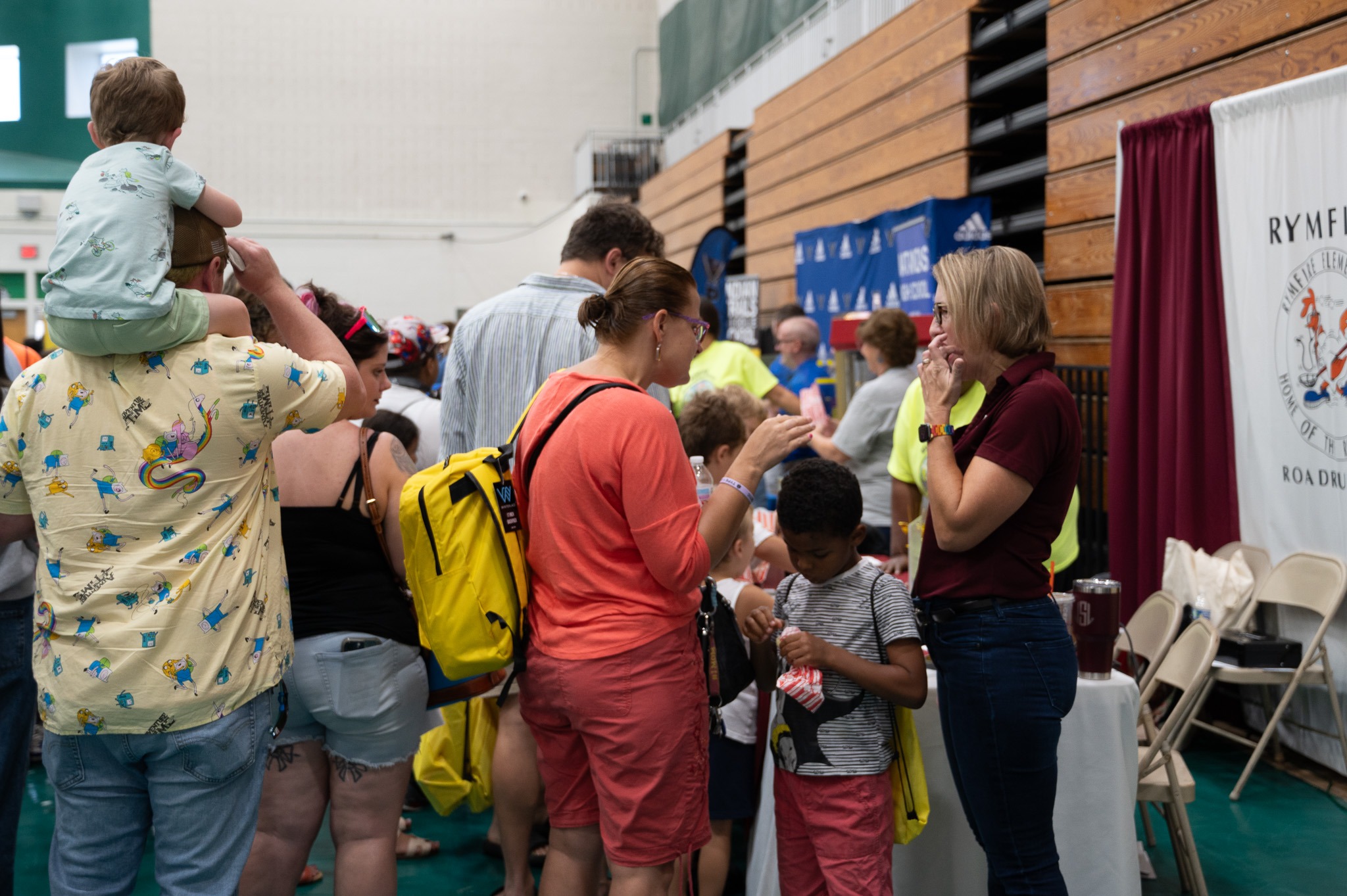 A crowd of people, including children, gather at various booths inside a gymnasium for a community event Some wear backpacks and hold bags A woman in a maroon shirt engages with attendees near a booth displaying a white banner and a sign with the word "RYMF" visible