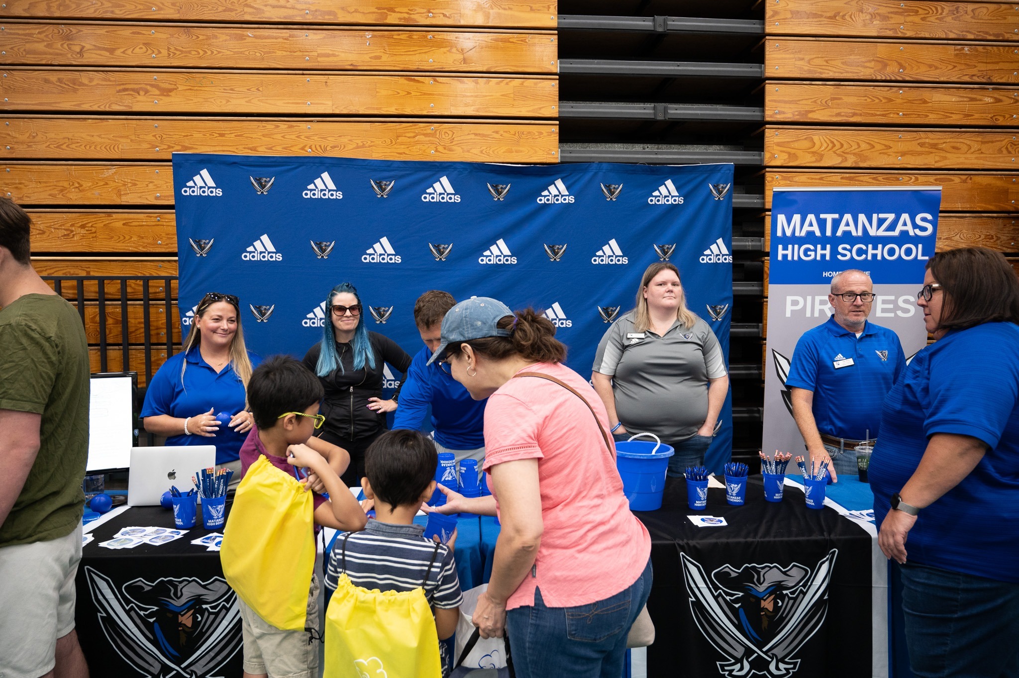 A group of people, including adults and children, are at a Matanzas High School booth in a gymnasium The booth has various items and is decorated with blue and white branding The backdrop displays the school's name and mascot Some people are interacting with each other