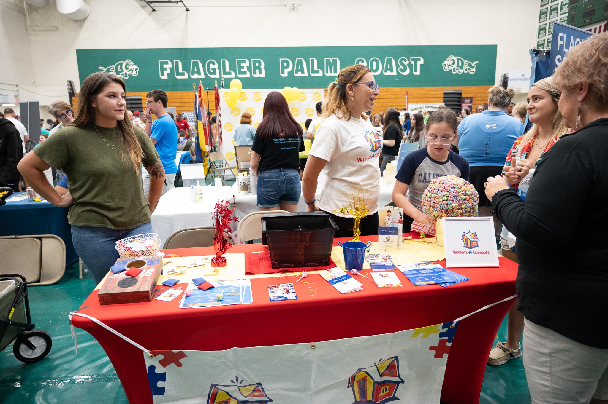 People stand near a table covered with educational materials and displays in a gymnasium with the banner "Flagler Palm Coast" in the background Various items, such as brochures and a gumball machine, are on the table The atmosphere is lively and social