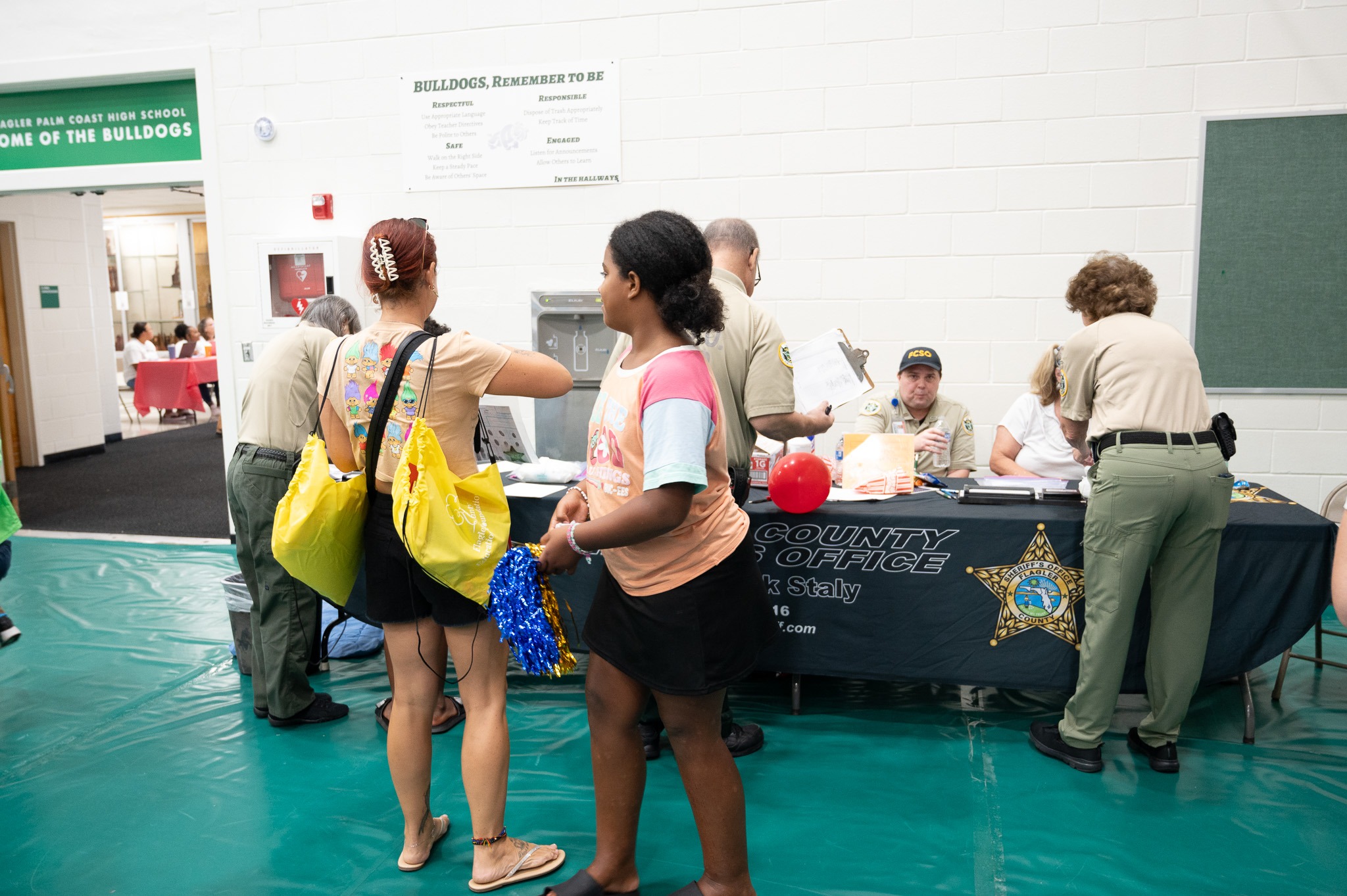 People interacting with law enforcement officers at a county sheriff's office booth in a gymnasium A girl stands in the foreground holding a blue pom pom Various informational materials are spread out on the table in the background