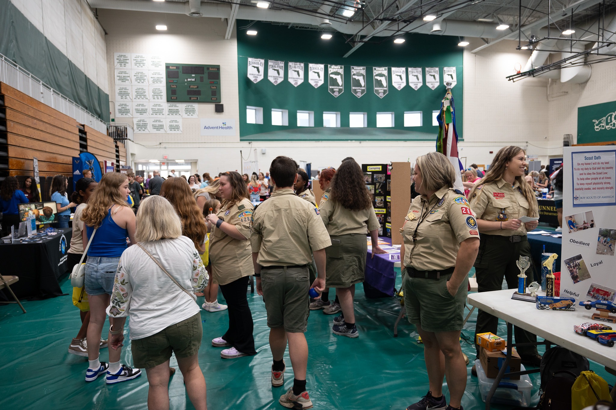 A group of people, including several uniformed Scouts, stand in a gymnasium that is hosting a fair or exhibition There are various booths and tables set up with displays and informational materials The gym is decorated with banners and flags