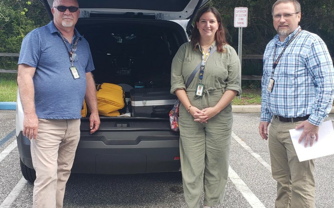 Three people stand next to an open trunk of a vehicle in a parking lot The trunk is filled with equipment The individuals, two men and a woman, are dressed casually and wear ID badges Trees are visible in the background