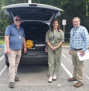 Three people stand next to an open trunk of a vehicle in a parking lot The trunk is filled with equipment The individuals, two men and a woman, are dressed casually and wear ID badges Trees are visible in the background