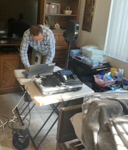 A person in a blue plaid shirt and khakis is working on a laptop placed on a small white table The room contains medical supplies, electronics, and a hospital bed Sunlight streams through a window with vertical blinds, casting shadows on the floor