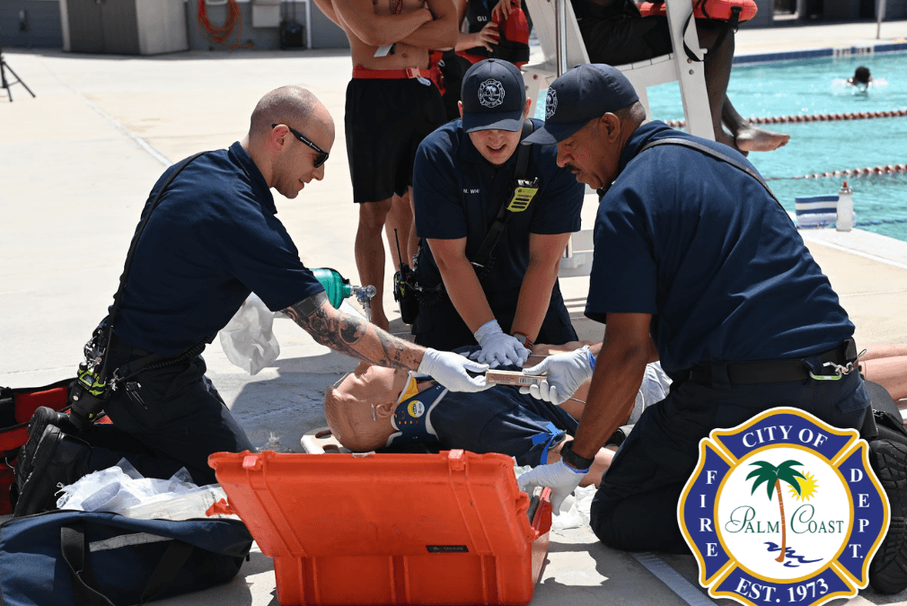 Three emergency responders are assisting a person lying on the ground beside a pool One responder is holding a piece of equipment, while the others are providing support Nearby, a fourth person observes, and a lifeguard is standing on the lifeguard chair