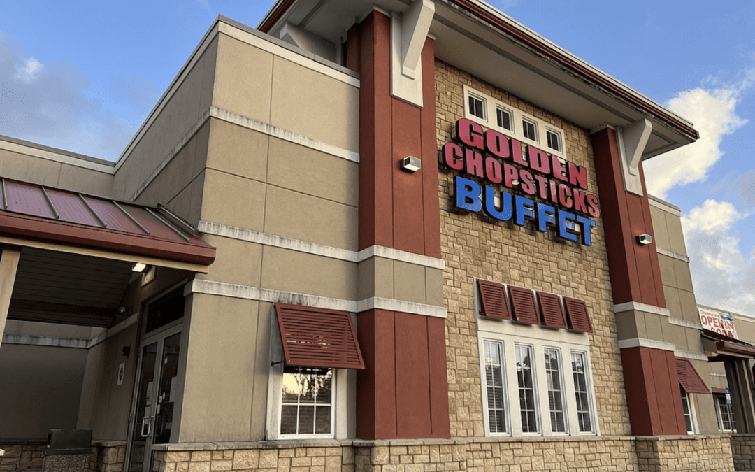 A restaurant with beige, tan, and red facade, featuring a sign that reads "Golden Chopsticks Buffet" in blue and red letters The entrance has large windows and a red awning The sky is partly cloudy