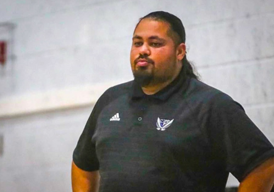 A man with long dark hair tied back, a beard, and a mustache is standing with his hands on his hips, wearing a black Adidas polo shirt with a sports team logo, against a plain, light colored wall He has a thoughtful or serious expression