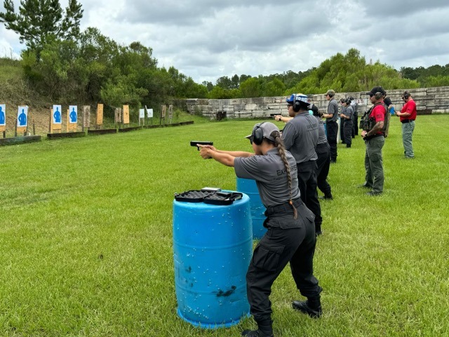 People participate in a handgun shooting practice at an outdoor firing range They are spaced out in a line, aiming at blue human silhouette targets mounted on stands The range is surrounded by grassy areas and trees, and the sky is cloudy