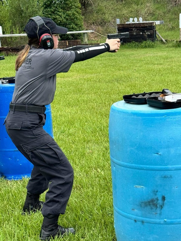 A person wearing a black cap, ear protection, and gray shirt with text on the back, practices shooting a handgun at an outdoor range Two blue barrels and a grassy field can be seen, with targets set up in the background