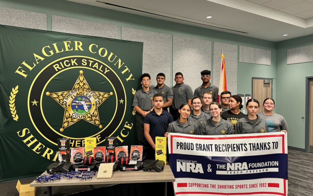 A group of people stand in front of the Flagler County Sheriff's banner They are holding and standing beside various items and a large sign that reads, "Proud grant recipients thanks to the NRA & The NRA Foundation," along with some promotional material