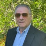 A man with gray hair wearing aviator sunglasses, a light blue collared shirt, and a dark blazer stands in front of lush green foliage He is looking at the camera with a slight smile