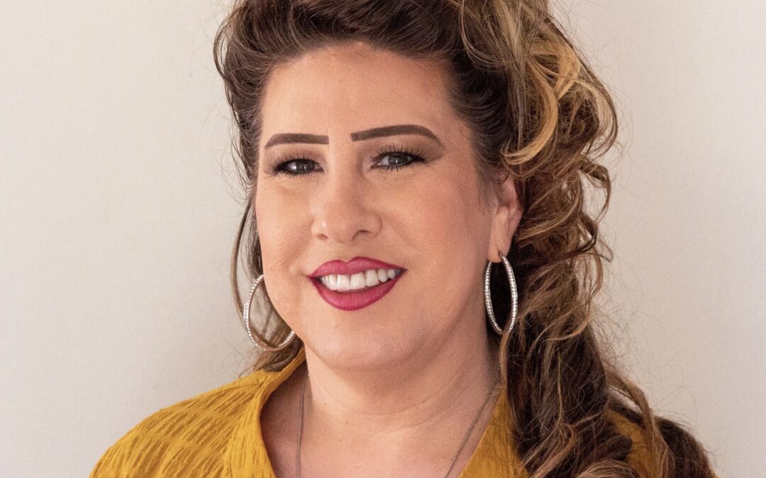 A woman with long wavy hair styled half up, wearing hoop earrings and a mustard yellow jacket over a black top, smiles while standing in front of a plain light colored background