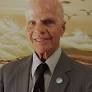 An elderly man, with short white hair, wearing a dark grey suit, white dress shirt, and patterned tie, smiles at the camera The background features a painting with light, airy colors, possibly depicting a landscape or abstract scene