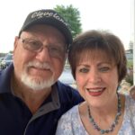 An elderly man wearing glasses, a Cleveland cap, and a polo shirt, smiles next to a woman with short brown hair, wearing a blue floral patterned blouse and a blue necklace They appear to be outdoors on a sunny day