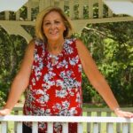 A woman with blonde hair stands smiling in a white wooden gazebo wearing a red, sleeveless, floral blouse She holds onto the gazebo railings with both hands The background is lush with green trees and foliage, suggesting an outdoor garden setting