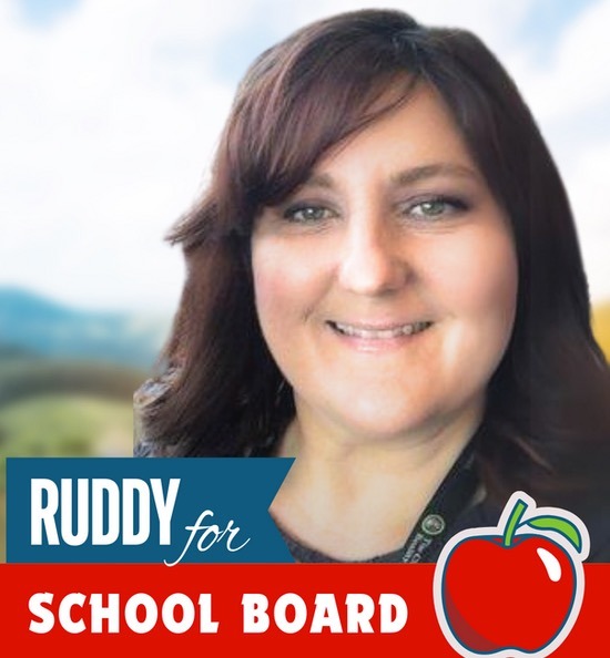 A woman with dark brown hair and a friendly smile stands in front of a blurred outdoor background There is a blue banner with the text "RUDDY for" and a red banner with the text "SCHOOL BOARD" alongside an illustration of a red apple