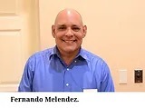 A smiling man with a shaved head wearing a blue collared shirt stands in front of a beige background There is text at the bottom of the image that says "Fernando Melendez