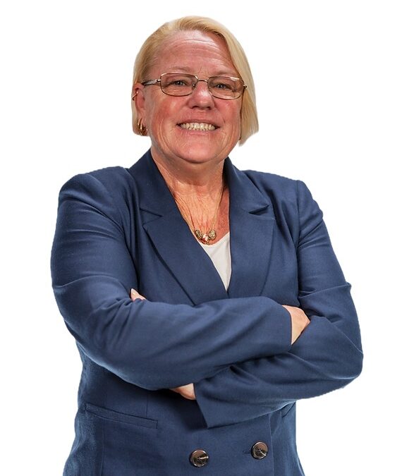 A woman with short blonde hair and glasses stands confidently with her arms crossed She is wearing a navy blue blazer and a white top, smiling at the camera against a plain white background