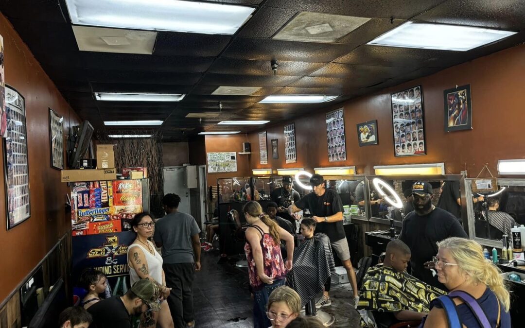 A busy barbershop with customers waiting and getting haircuts The shop has several barbers working on clients, while other patrons wait and chat The walls are adorned with posters and haircut style charts The space is brightly lit with overhead lights