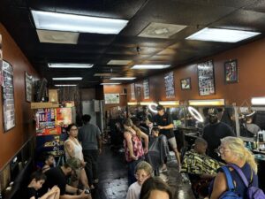 A busy barbershop with customers waiting and getting haircuts The shop has several barbers working on clients, while other patrons wait and chat The walls are adorned with posters and haircut style charts The space is brightly lit with overhead lights