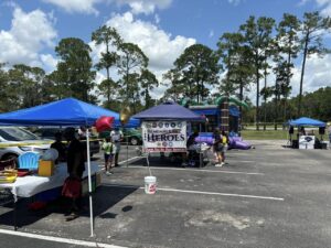 A community event in a parking lot features tents and booths under a sunny sky People mingle and browse at various setups, including a tent with a "Remember Our Heroes" sign Inflatable structures and tall trees are visible in the background