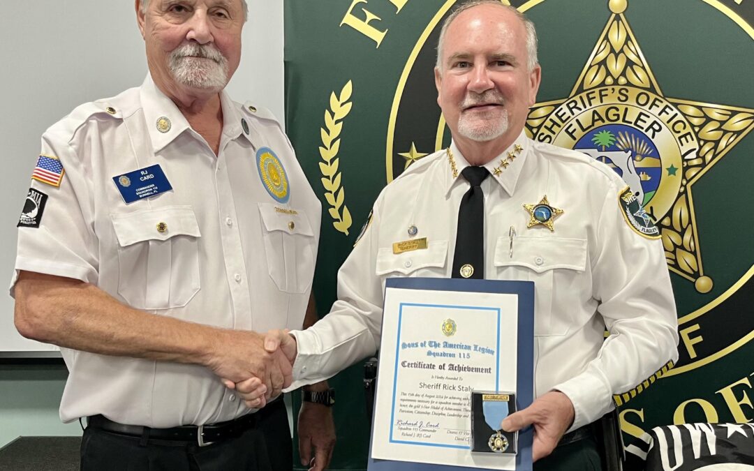 Two uniformed men shaking hands, one presenting a certificate and a badge to the other The recipient, wearing a sheriff's uniform, stands in front of a backdrop with a sheriff's office badge and the name Rick Staly Both are smiling in a formal setting