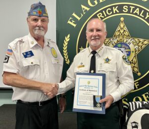 Two uniformed men shaking hands, one presenting a certificate and a badge to the other The recipient, wearing a sheriff's uniform, stands in front of a backdrop with a sheriff's office badge and the name Rick Staly Both are smiling in a formal setting