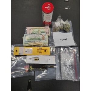 A table displays several items including a can of Coca Cola, a stack of assorted currency, two small bags of green plant material, a plastic pouch with a gray powdery substance, three packaged vape cartridges, a knife, and a card with the brand "Kynd