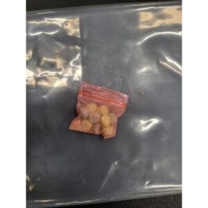 A small red ziplock bag containing several circular, yellowish orange tablets is placed on a larger, transparent, and slightly reflective surface The background appears dark and slightly textured