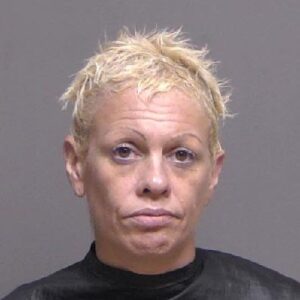 A woman with short, spiky blond hair and a serious expression is photographed against a plain gray background She is wearing a dark colored top