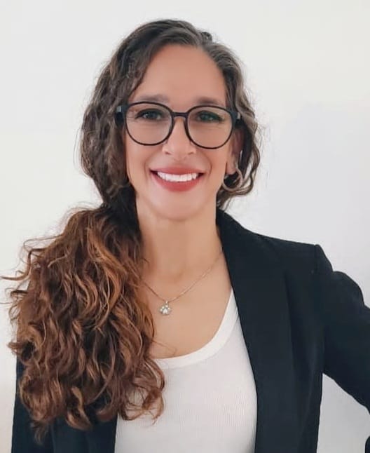 A woman with long, curly brown hair and glasses is smiling She is wearing a black blazer over a white top and a necklace with a small, round pendant The background is plain white