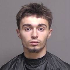 A young man with short, messy hair and a trimmed goatee is wearing a black cape typically used during haircuts He is facing the camera with a neutral expression, set against a plain grey background