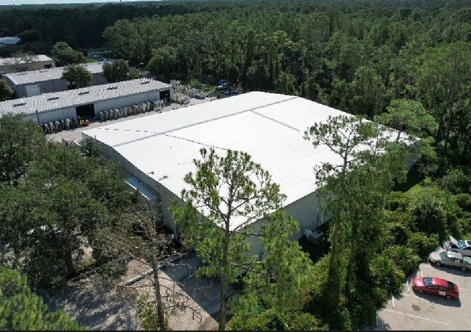 Aerial view of a large, white roofed industrial warehouse surrounded by dense green trees Several smaller buildings are situated nearby, with a few parked vehicles visible The scene includes parking areas and a backdrop of forested area