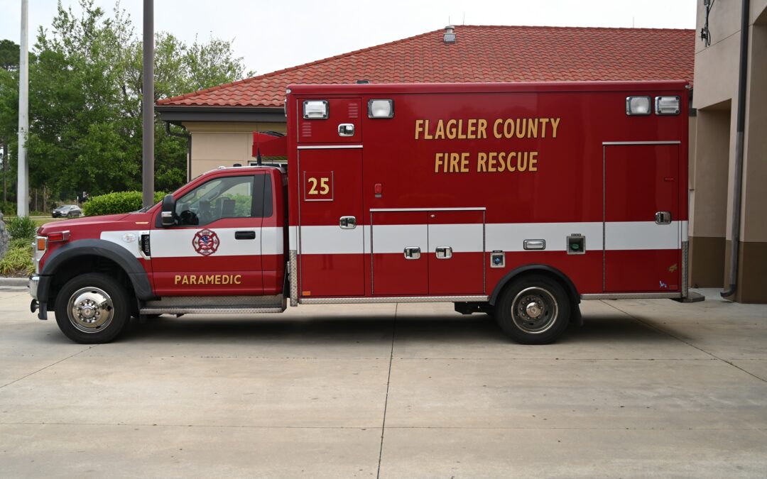 A red emergency medical service truck with "Flagler County Fire Rescue" and "" written on the side is parked outside a building The truck has white and red stripes, a paramedic emblem, and various compartments for equipment