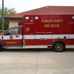 A red emergency medical service truck with "Flagler County Fire Rescue" and "" written on the side is parked outside a building The truck has white and red stripes, a paramedic emblem, and various compartments for equipment